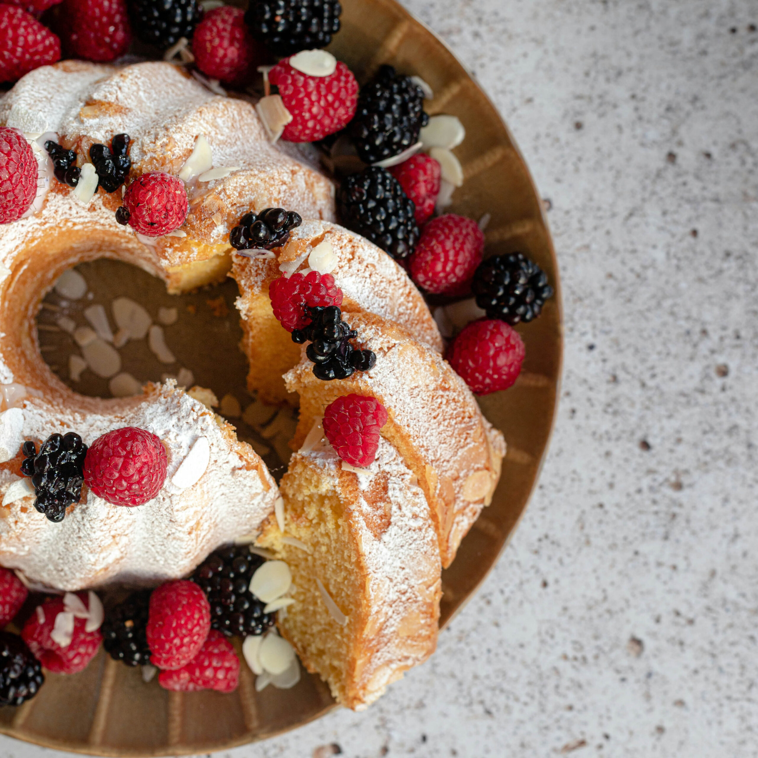 Gugelhupf mit Staubzucker, Himbeeren, Brombeeren und Mandelblättchen auf goldenem Teller
