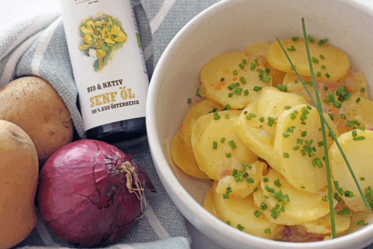 Steirischer Kartoffelsalat mit Schnittlauch in Schüssel, daneben Schalk Mühle Senföl, Erdäpfel und rote Zwiebel