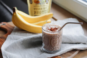 Schoko-Bananen-Smoothie im Glas mit Kakao und Metallstrohhalm, dahinter Bananen und Schalk Mühle Vegan High Protein