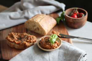 Roter Gemüseaufstrich auf einer Brotscheibe und im Schälchen mit Basilikum, dahinter ein Weißbrotlaib und Tomaten