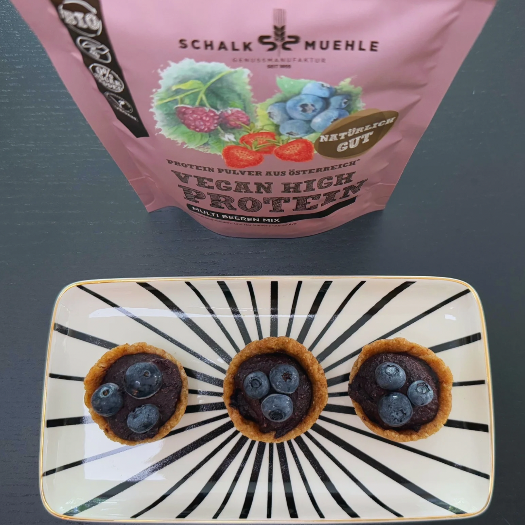 Drei Mini-Schoko-Törtchen mit Heidelbeeren neben Schalk Mühle Vegan High Protein Multi Beeren Mix