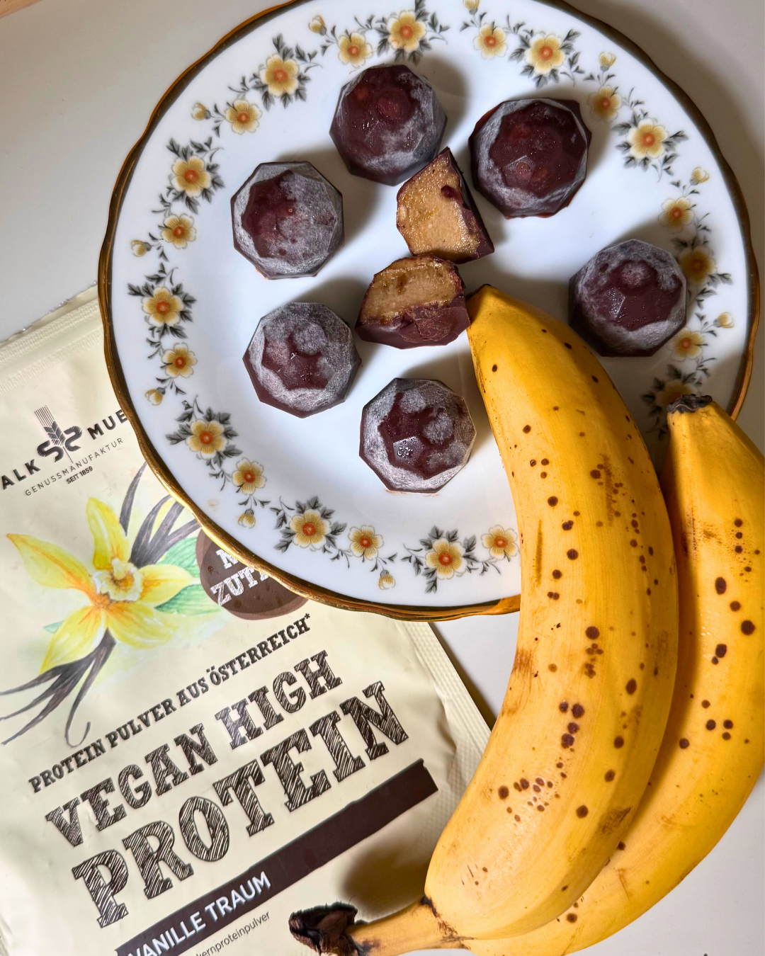 Schokolierte Bananenstücke auf einem Blumenteller mit Bananen neben Schalk Mühle Vegan High Protein Vanille Traum