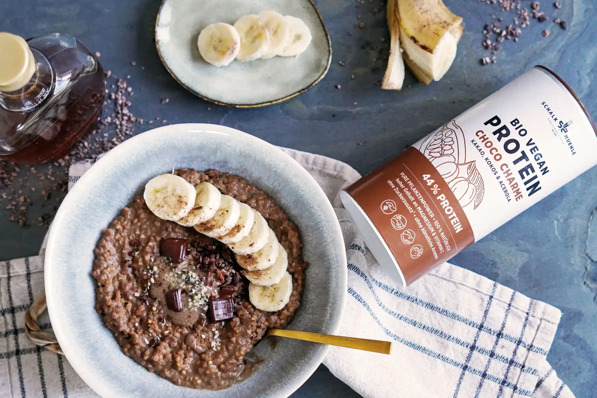 Chocolate porridge with banana slices, chocolate pieces and hemp seeds, next to Schalk Mühle Organic Vegan Protein Choco Charme