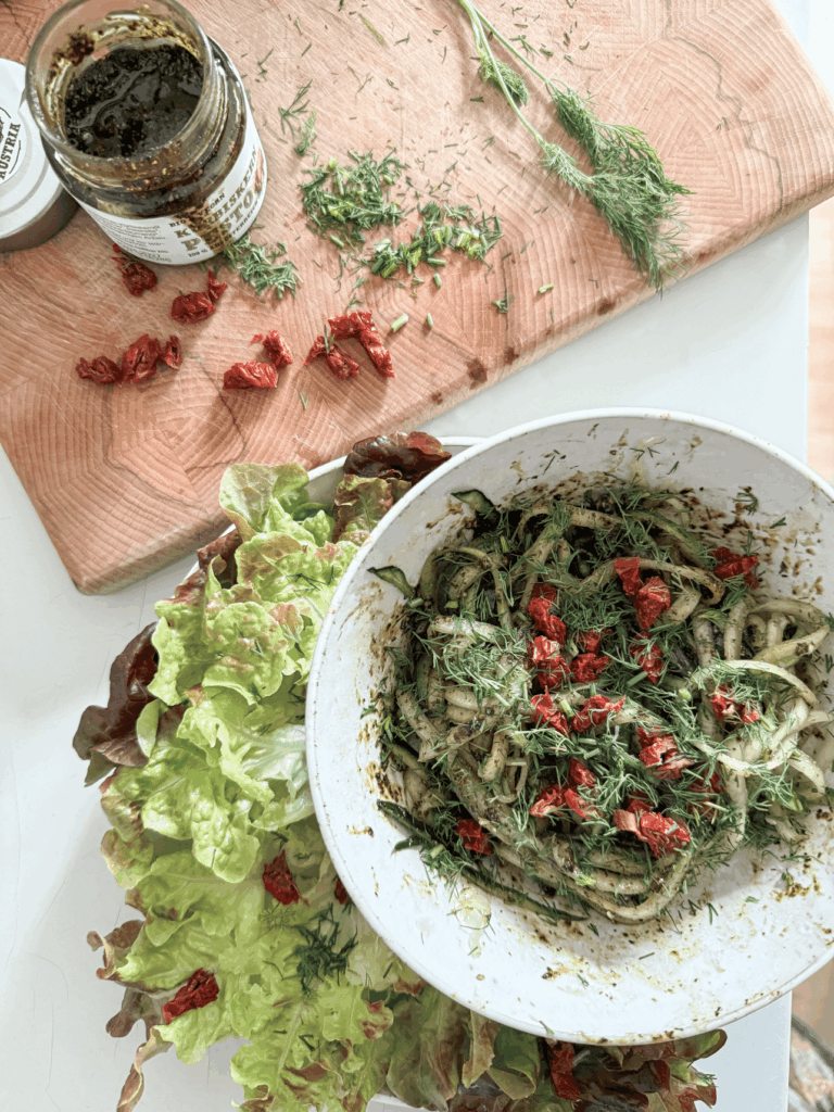 Sommerliche Gurken Spaghetti mit Schalk Mühle Bio Kürbiskern Pesto, angerichtet mit frischem Salat und Gewürzen in einer weißen Schüssel auf einem Holzbrett.