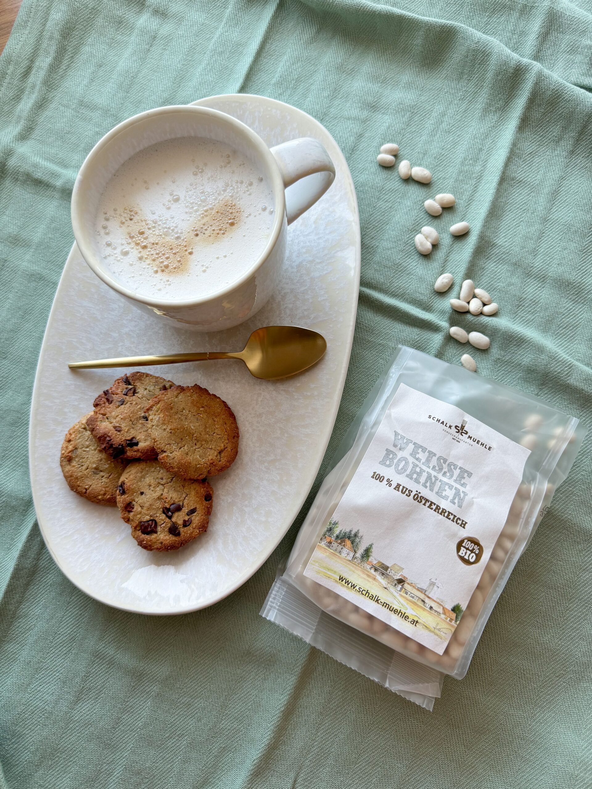Weiße-Bohnen-Cookies mit Schokostückchen und Milchkaffee, daneben Schalk Mühle Weiße Bohnen