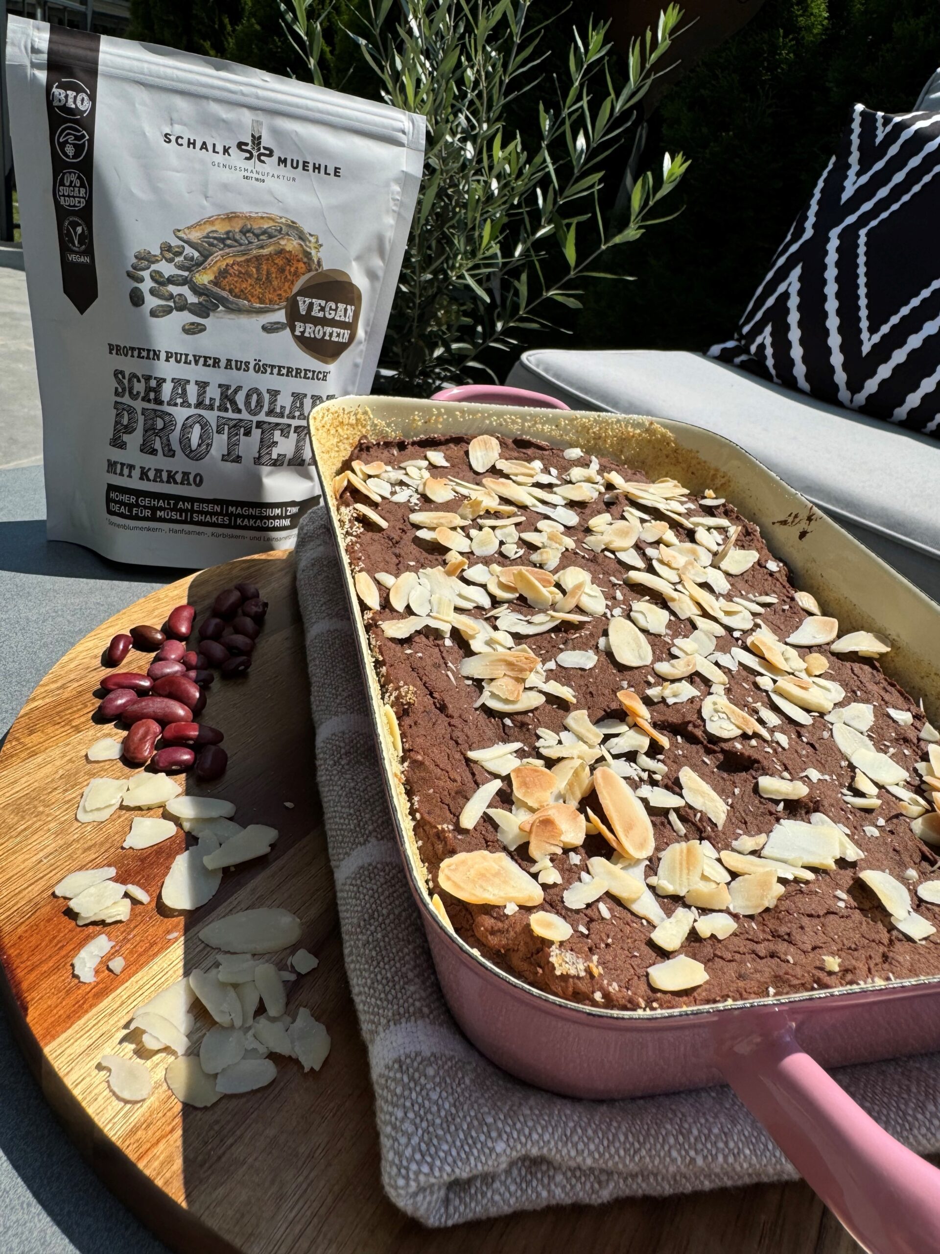 Veganer Schoko-Bohnenkuchen mit Mandelblättchen in der Form, daneben Schalk Mühle Protein mit Kakao