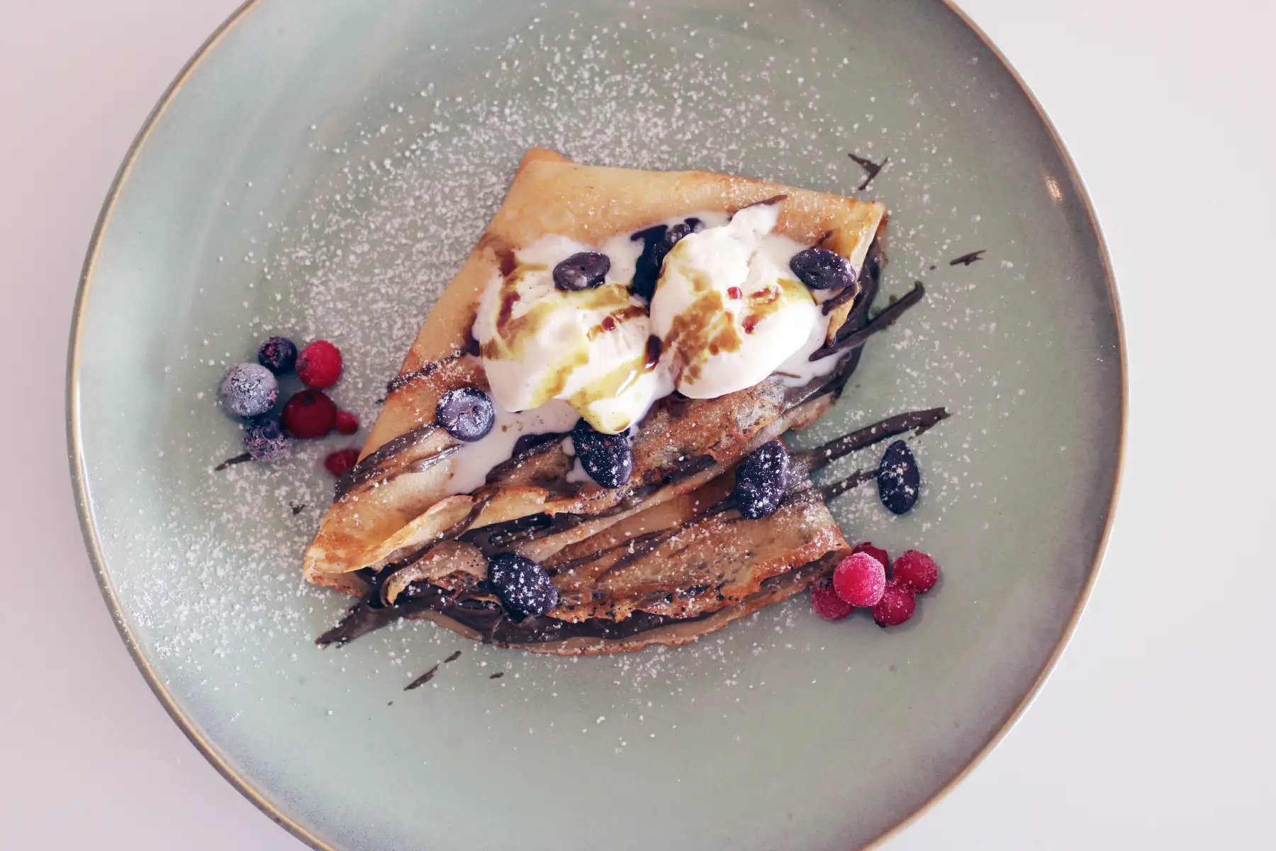 Filled ice crêpe with chocolate sauce, vanilla ice cream, berries and icing sugar on a green plate