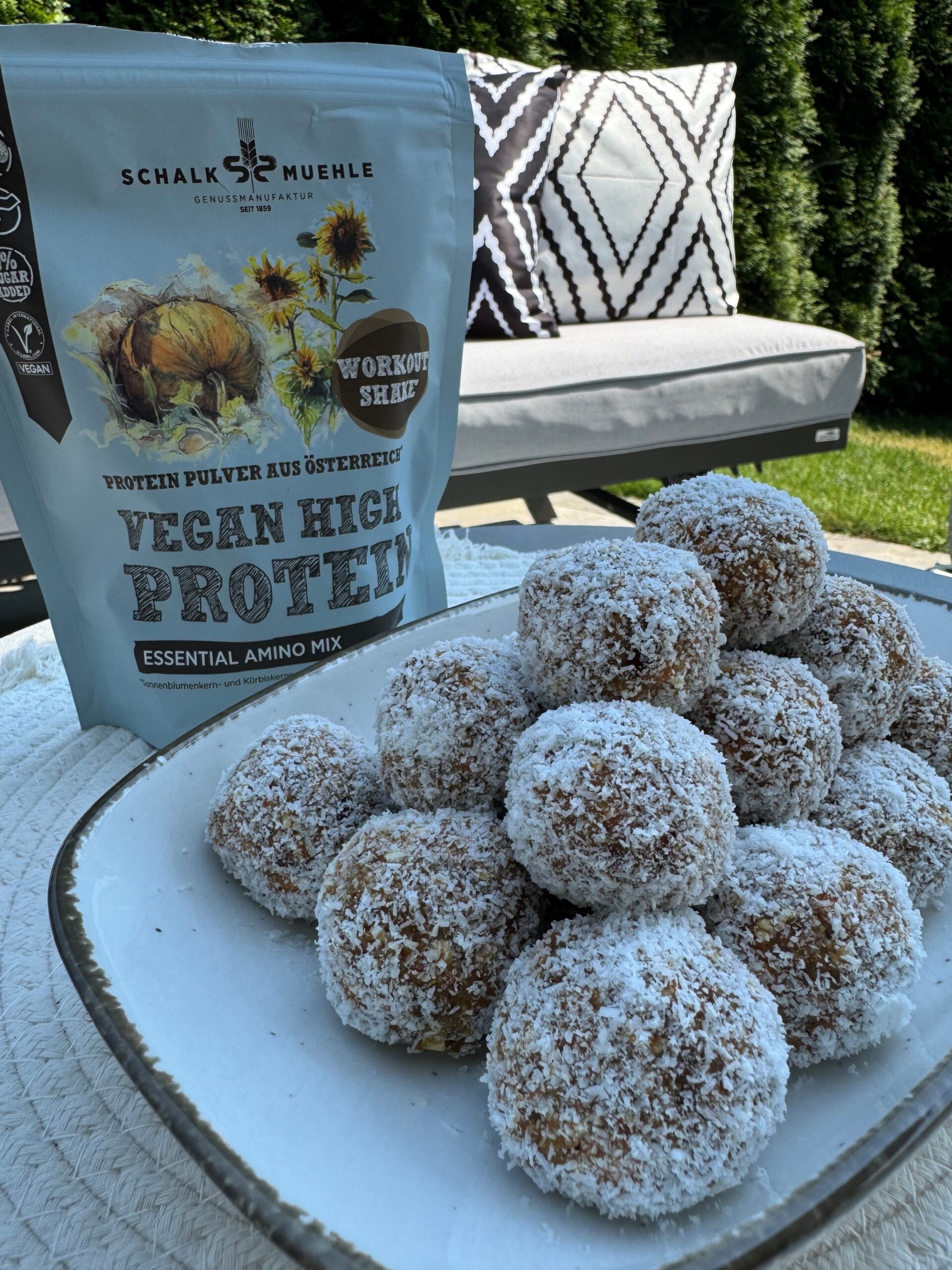 Vegane Kokos-Energiekugeln auf Teller im Garten, daneben Schalk Mühle Vegan High Protein