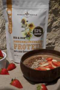 Smoothie-Bowl in Kokosschale mit Erdbeeren und Banane, dazu Schalk Mühle Sonnenblumenkern-Protein