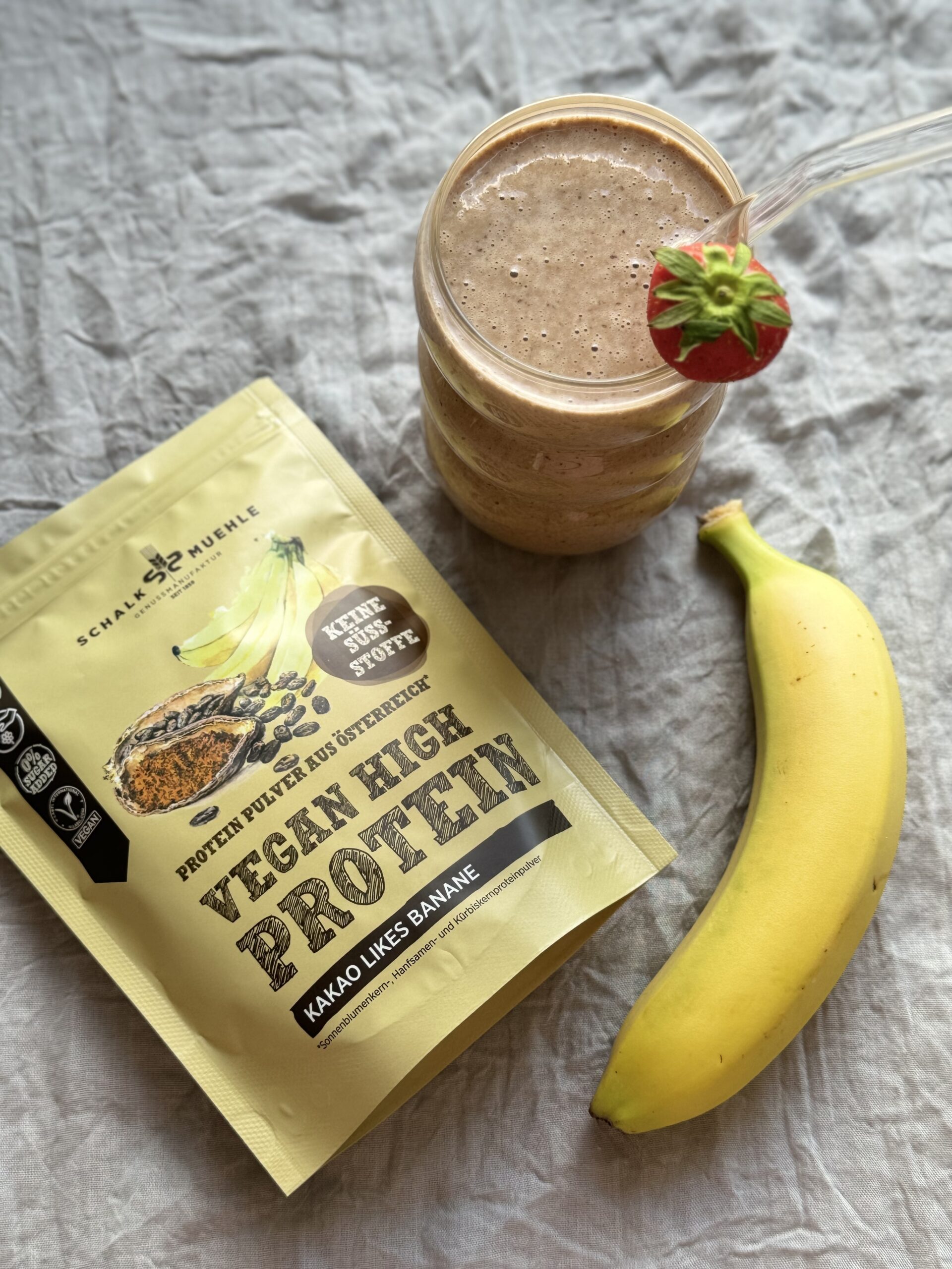 Frühstücks-Shake im Glas mit Erdbeere und Banane, daneben Schalk Mühle Vegan High Protein Kakao-Banane