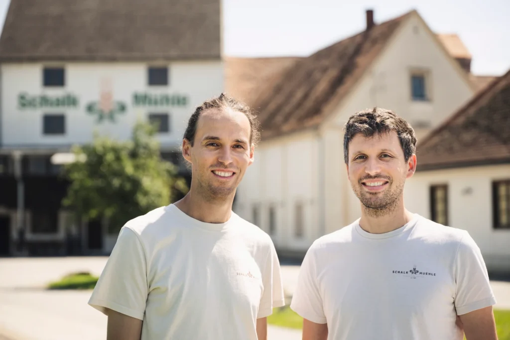 The two smiling managing directors of Schalk Mühle in white company T-shirts in front of the company building