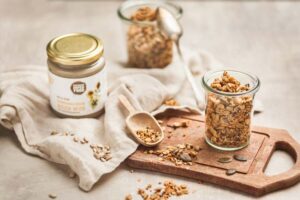 Knuspermüsli in Weckgläsern auf Holzbrett, daneben Glas Schalk Mühle Sonnenblumenkernmus und verstreute Kerne