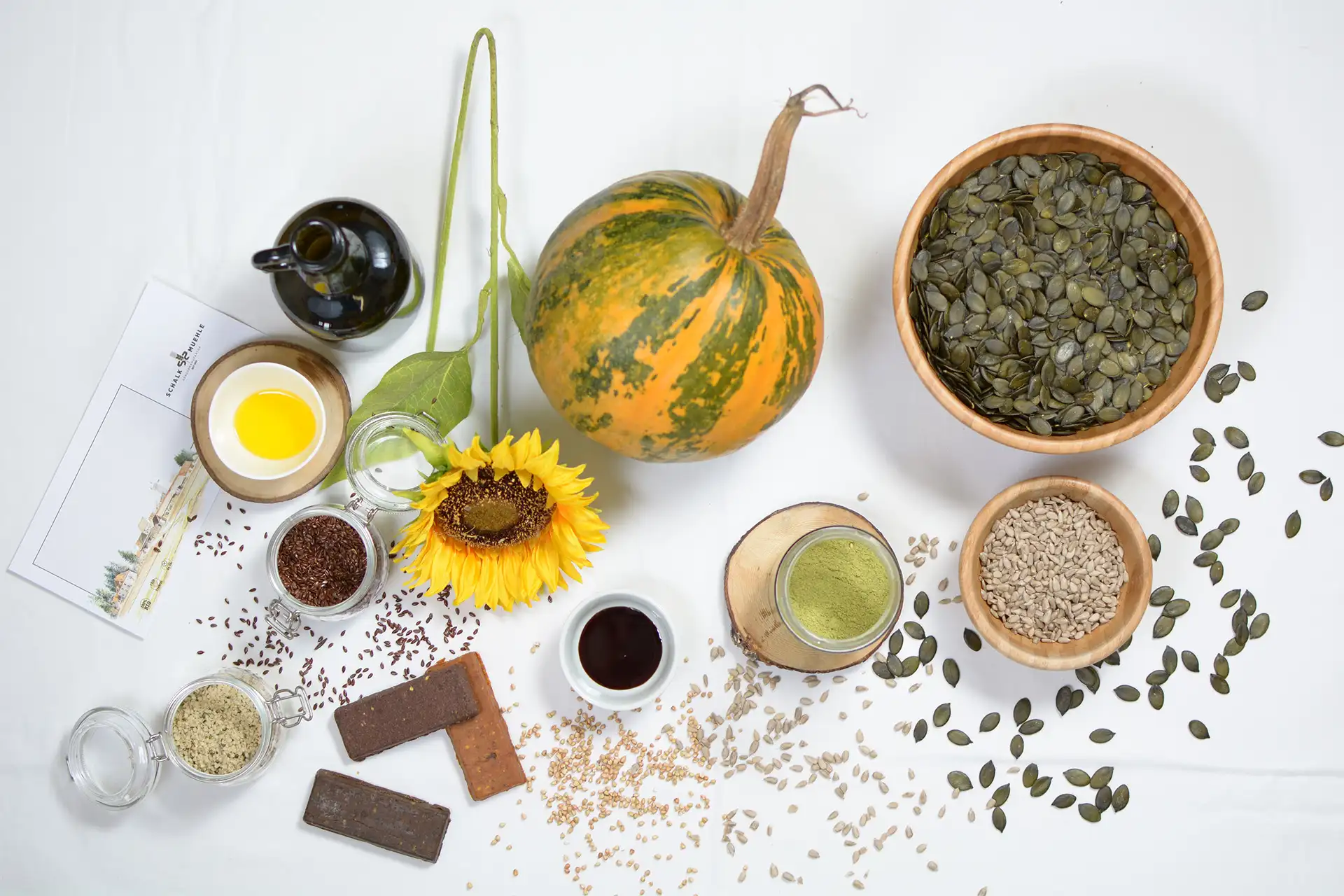 Ganzer Ölkürbis mit einer Schüssel Kürbiskerne, dazu Sonnenblumenkerne, Leinsamen und Kürbiskernöl der Schalk Mühle