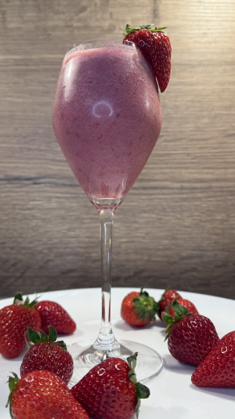 Rosafarbener Erdbeer-Smoothie im Stielglas, umgeben von frischen Erdbeeren