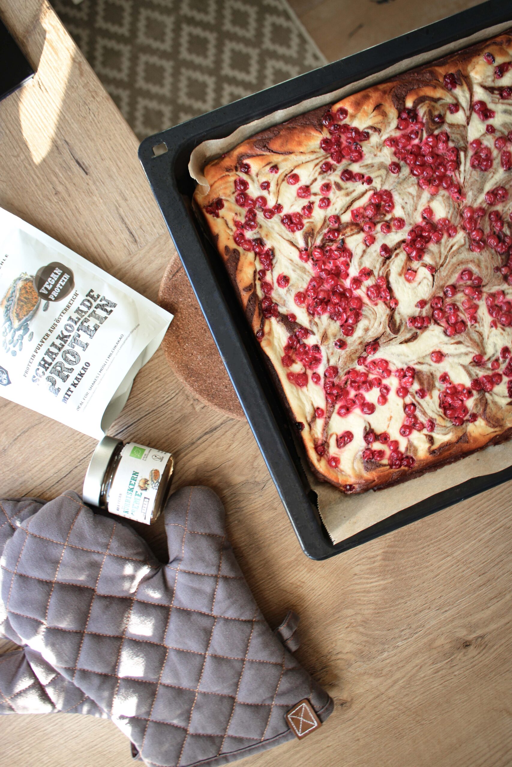 Marmorierter Topfenkuchen mit roten Ribiseln auf dem Blech, daneben Schalk Mühle Schokolade-Protein und Kürbiskerncreme