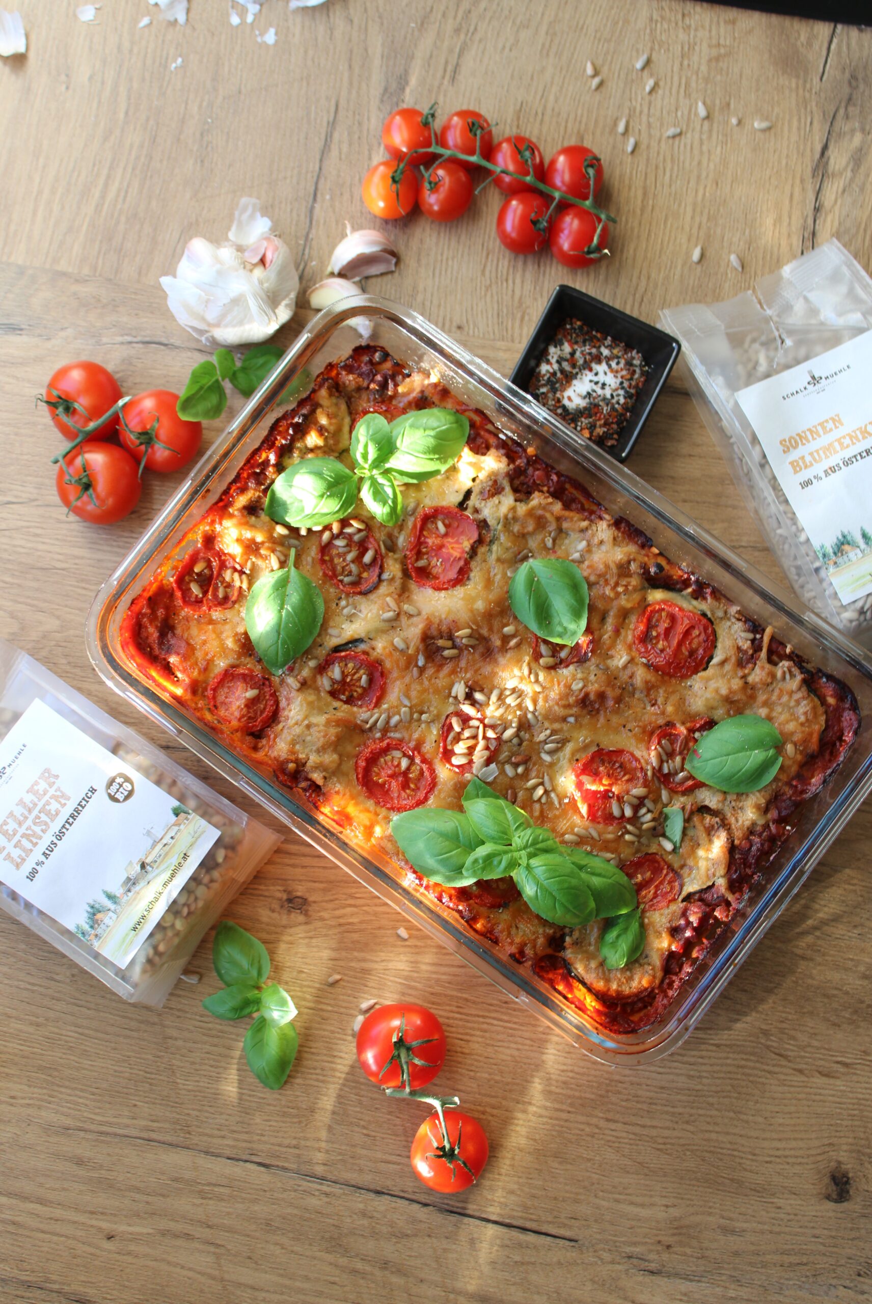 Gemüseauflauf mit Tomaten, Basilikum und Sonnenblumenkernen in Glasform, daneben Schalk Mühle Tellerlinsen und Sonnenblumenkerne