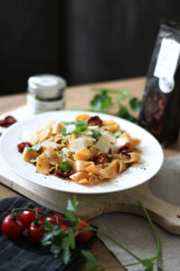Pappardelle mit getrockneten Tomaten, Parmesanspänen und Petersilie auf weißem Teller, daneben Rispentomaten
