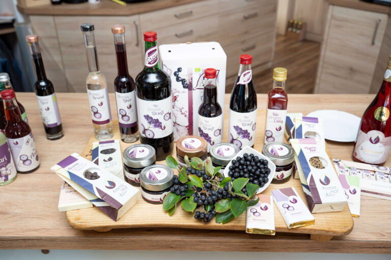 Eine Produktpräsentation von Aronia-Hof Kober auf einem Holzbrett mit verschiedenen Aroniabeeren-Produkten wie Säften, Likören, Schokolade, Tee und frischen Beeren in einer Küche.