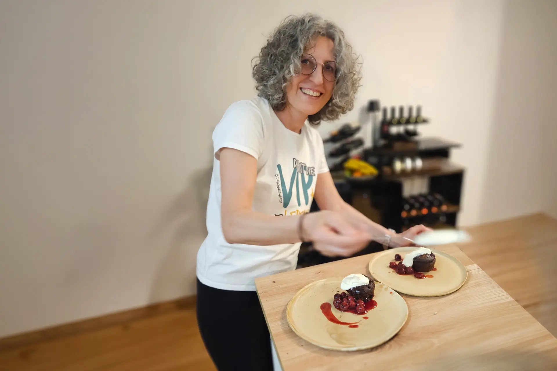 Lächelnde Frau richtet beim Kochworkshop zwei Teller mit Schokoküchlein, Kirschen und Sahne an