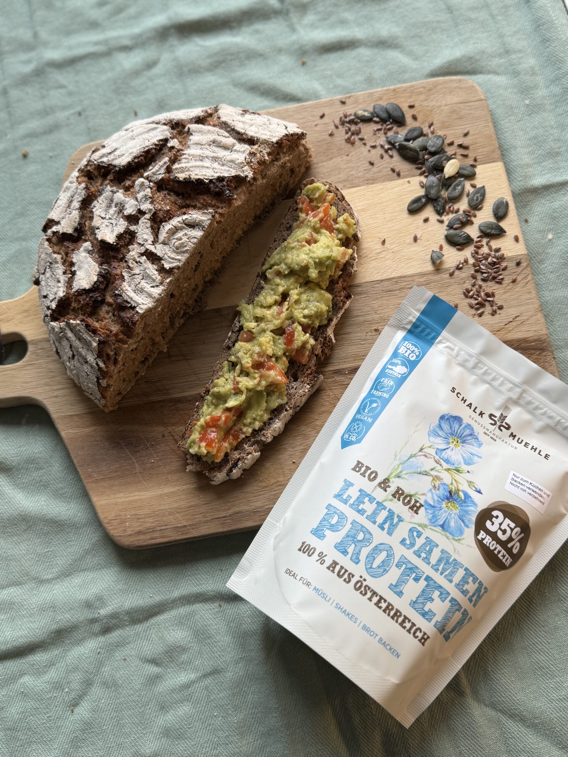 Dunkles Brot mit Avocado-Aufstrich auf Holzbrett, daneben Schalk Mühle Leinsamen Protein