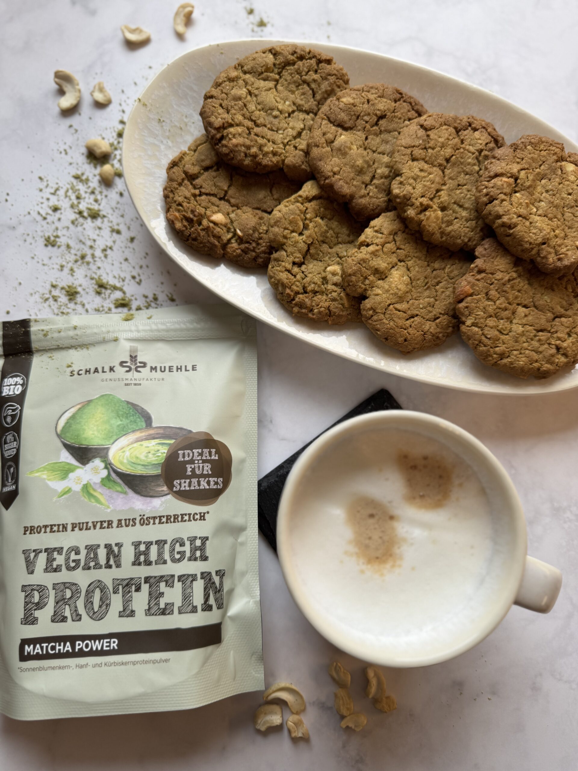 Matcha-Cookies auf Teller mit Schalk Mühle Matcha-Proteinpulver, Cappuccino und Cashewkernen