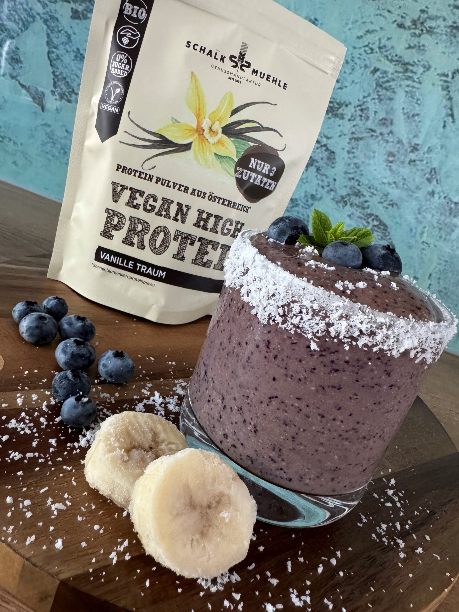 Blaubeer-Bananen-Smoothie im Glas mit Kokosrand, daneben Schalk Mühle Vegan High Protein Vanille