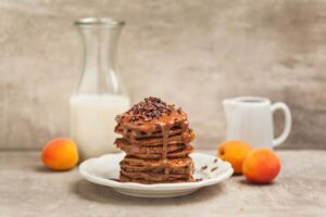 Stapel Schoko-Pancakes mit Karamellsauce und Kakaonibs, dazu Milchflasche und frische Marillen