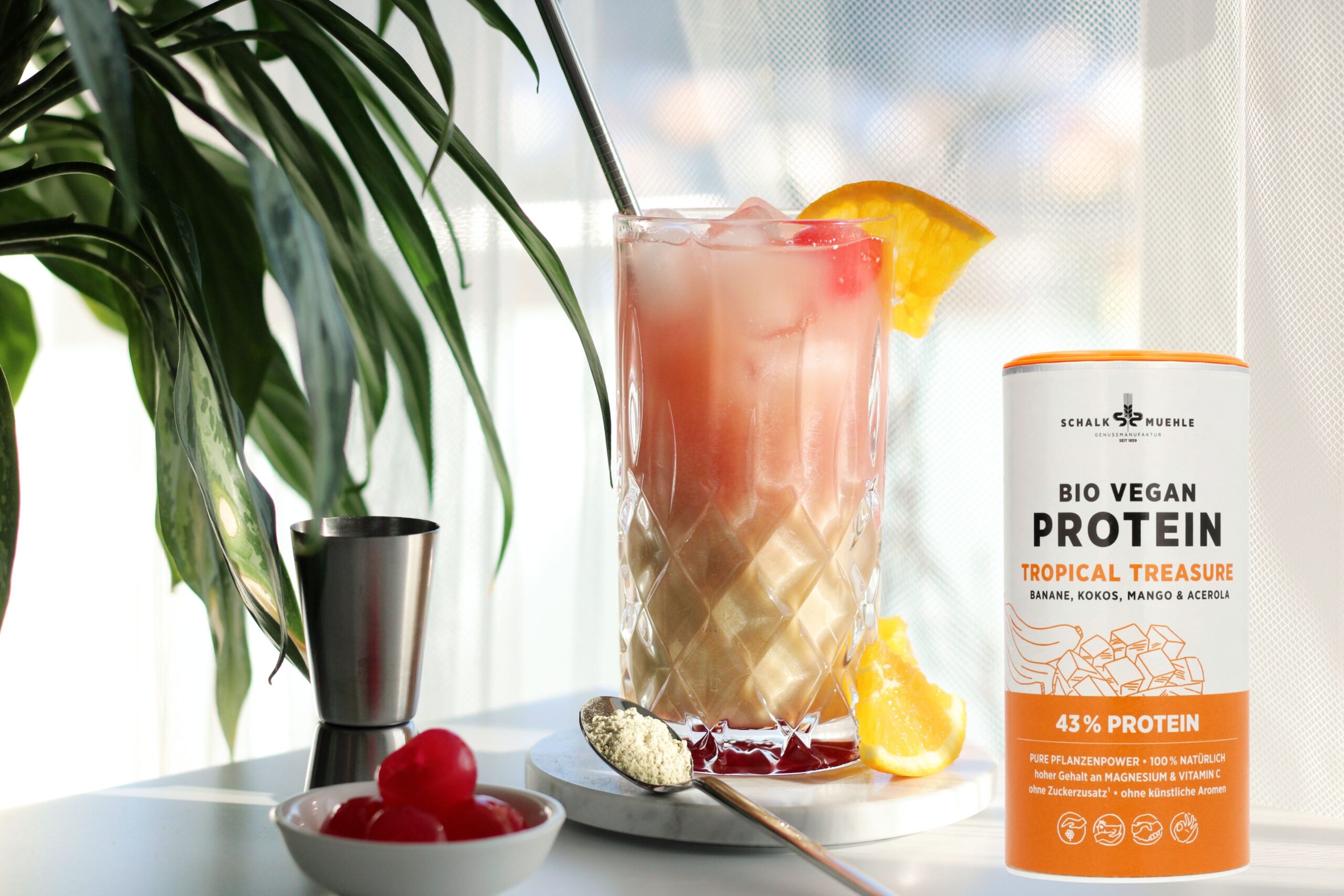 Fruchtiger Drink im Glas mit Orangenscheibe und Cocktailkirsche neben Schalk Mühle Bio Vegan Protein Tropical Treasure