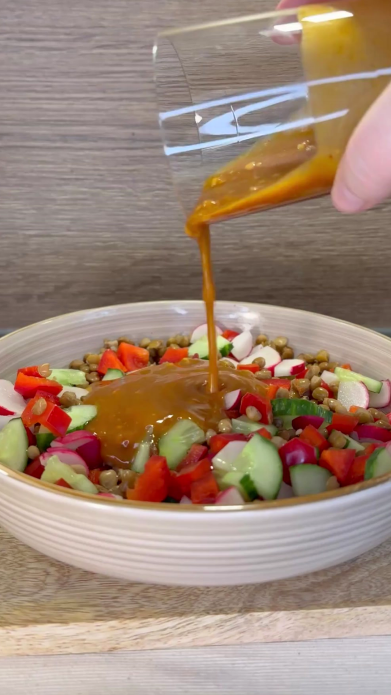 Hand gießt Dressing über bunten Linsensalat mit Gurke, Radieschen und Paprika