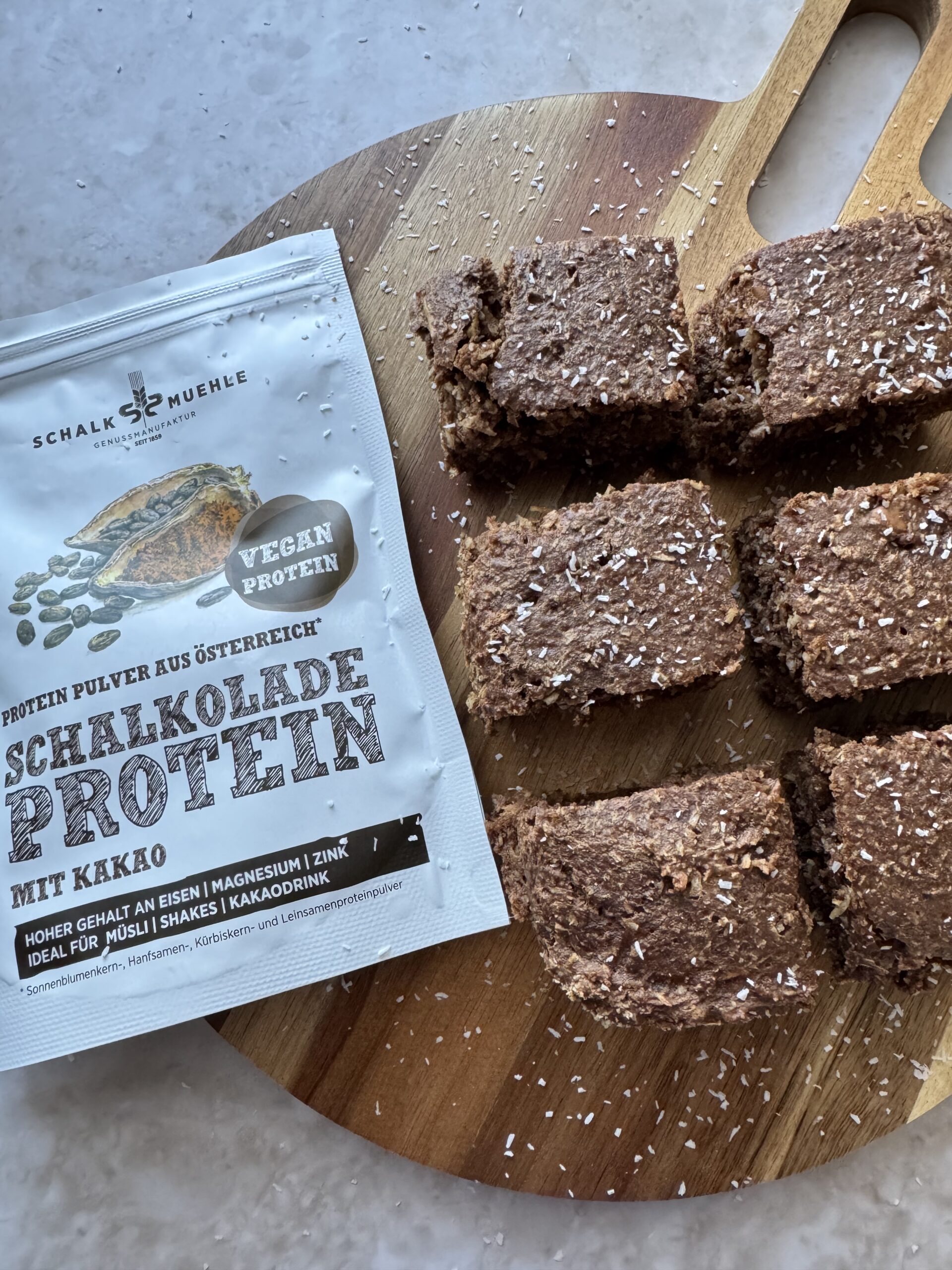 Schoko-Kokos-Kuchenstücke auf Holzbrett mit Kokosraspeln und Schalk Mühle Schalkolade-Proteinpulver