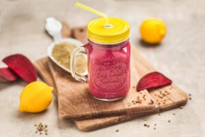 Pinker Smoothie im Bügelglas mit gelbem Deckel und Strohhalm, daneben rote Bete, Zitronen und Hanfsamen