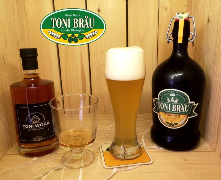 Ein Foto, das ein hohes Weizenglas mit Bier und Schaum, eine dunkle Bügelflasche Weizenbier, eine Flasche Whisky und ein teilgefülltes Whiskyglas auf Holzbrettern zeigt. Ein ovales grünes Logo mit dem Text "TONI BRÄU" hängt über den Getränken.