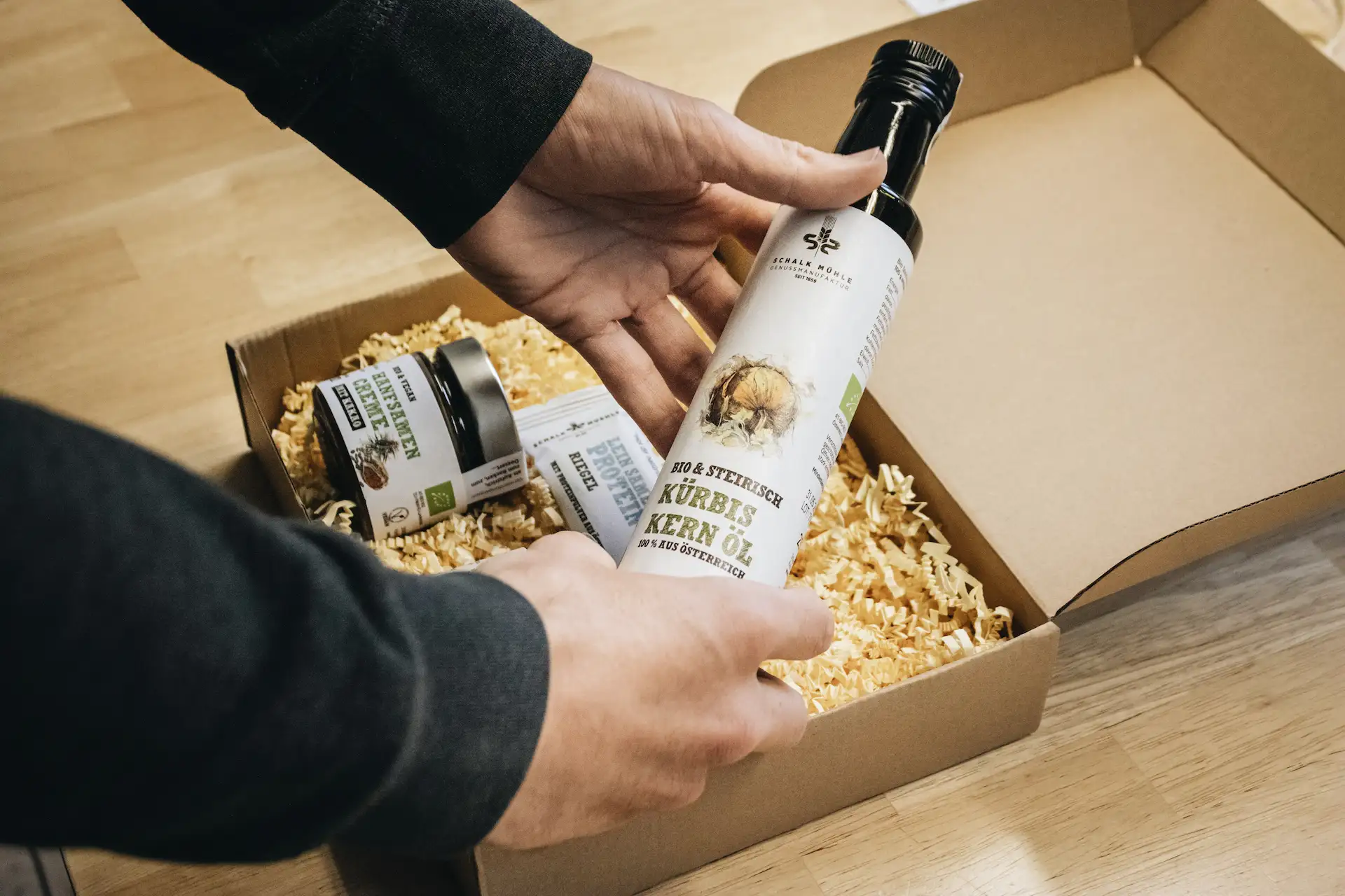 Hände legen eine Flasche Schalk Mühle Kürbiskernöl in ein mit Holzwolle befülltes Geschenkset mit weiteren Bio-Produkten