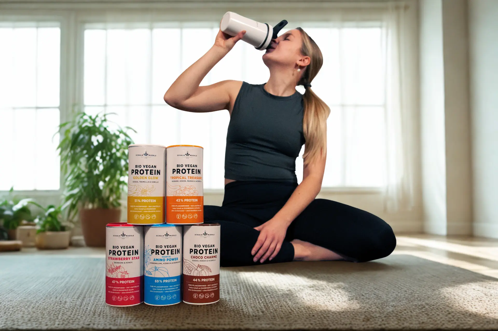 Frau trinkt einen Protein-Shake, davor fünf Schalk Mühle Bio Vegan Protein Dosen in bunten Sorten