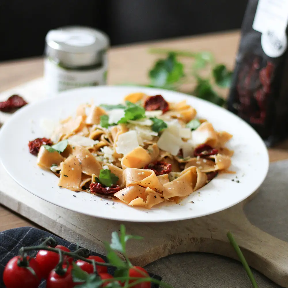 Teller Bandnudeln mit getrockneten Tomaten, Parmesan und Kräutern, dahinter ein Glas Schalk Mühle Pesto