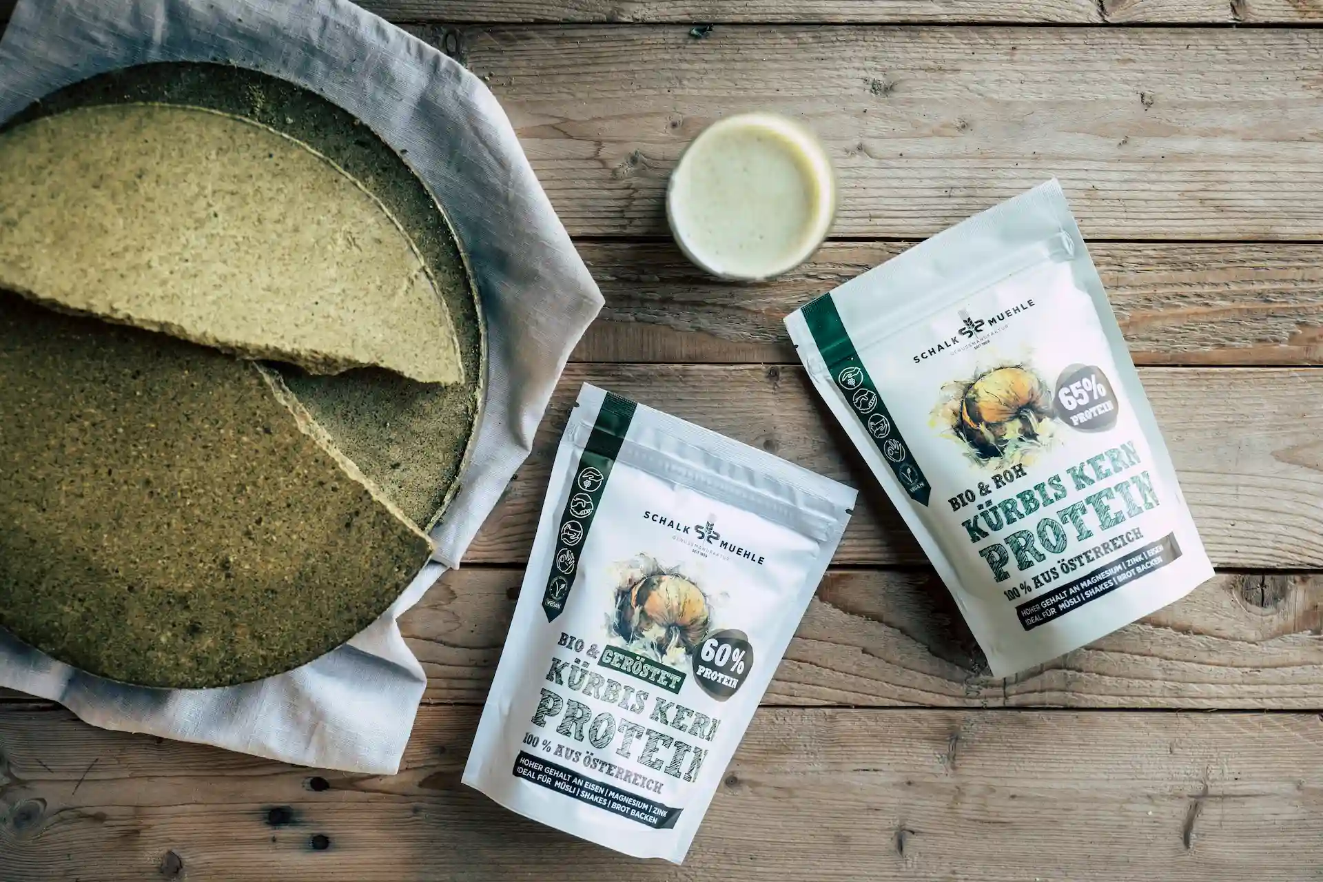 Zwei Schalk Mühle Kürbiskern Protein Pulver, geröstet und roh, neben einem Kürbiskern-Presskuchen