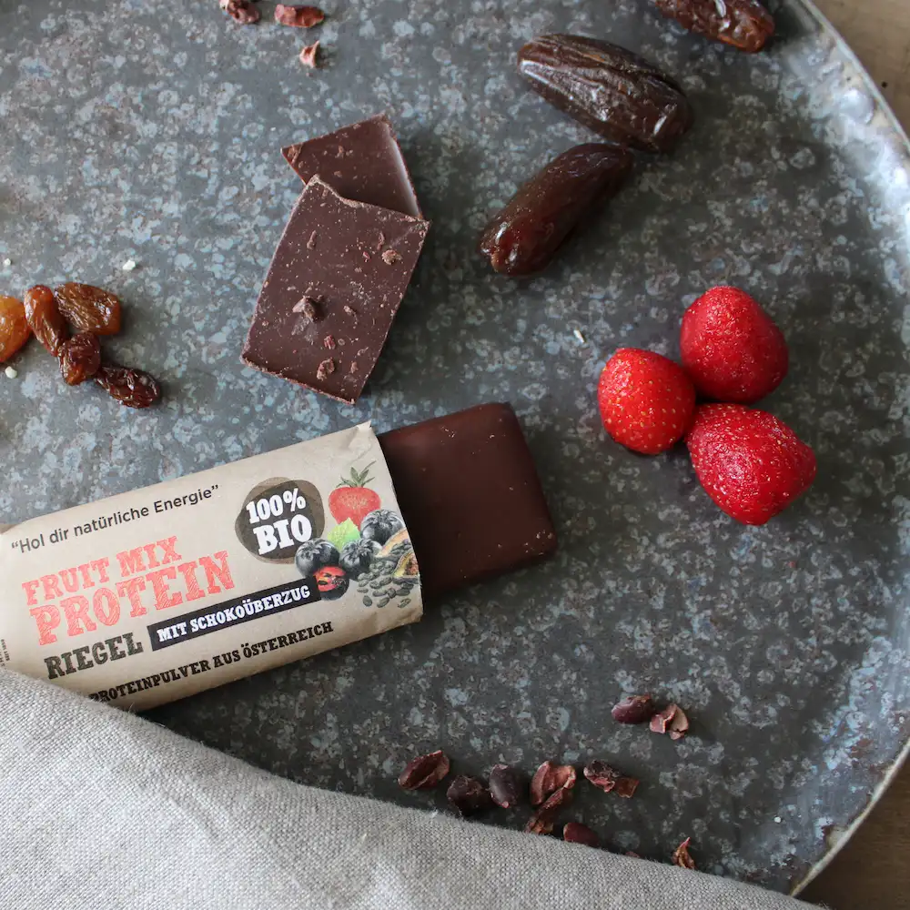 Angebissener Schalk Mühle Fruit Mix Protein-Riegel mit Schokoüberzug, dazu Erdbeeren, Datteln und Rosinen