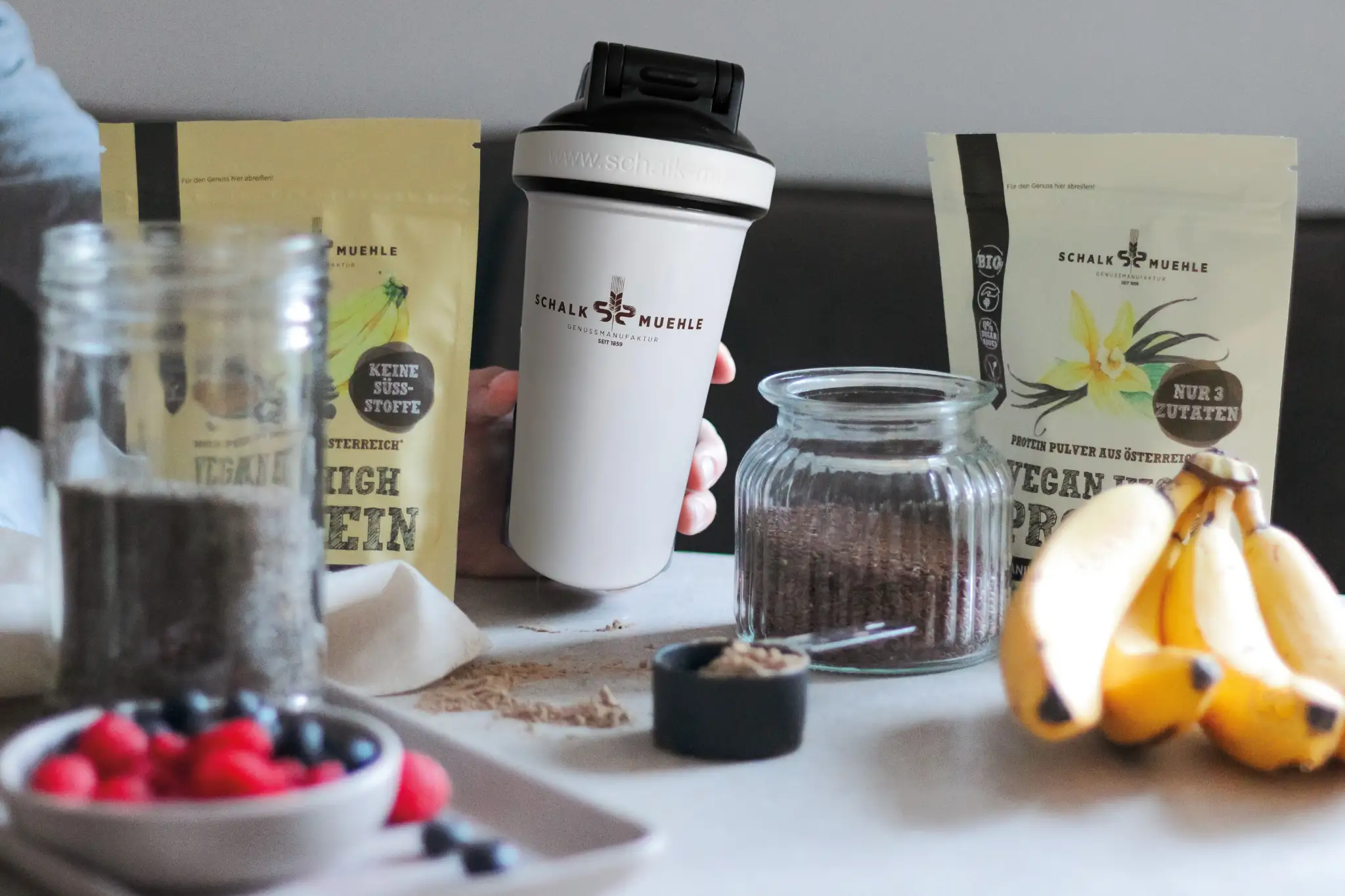 Weißer Schalk Mühle Shaker in der Hand, dahinter Protein-Pulver Sackerl, daneben Bananen und Beeren