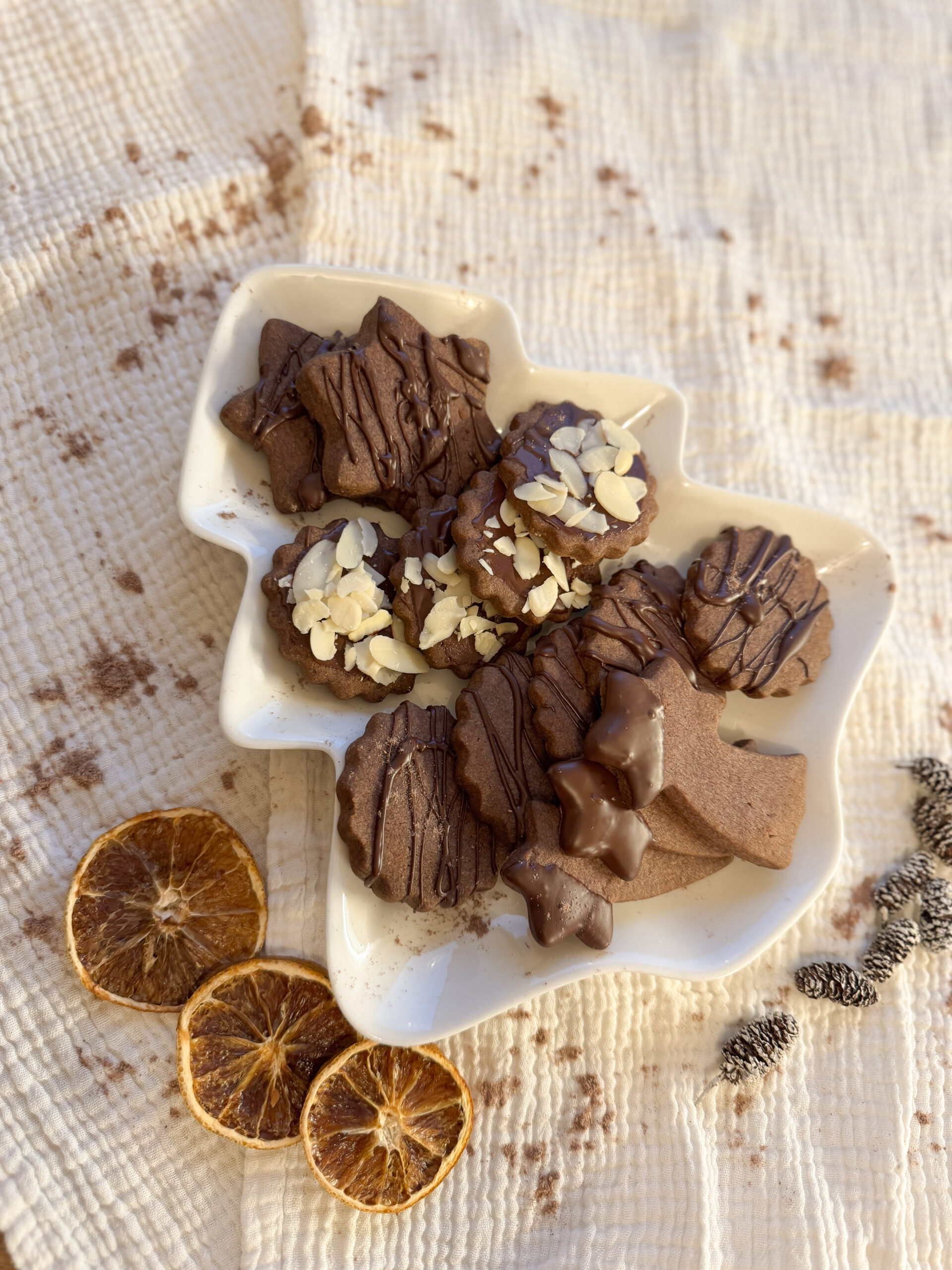 Schoko-Mürbteigkekse in Weihnachtsformen mit Schokoglasur und Mandeln, daneben getrocknete Orangenscheiben