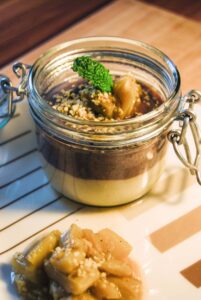 Zweierlei-Creme im Bügelglas mit Minzblatt, Hanfsamen und Fruchtkompott, daneben ein Löffel Kompott am Tellerrand