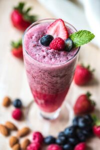 Beeren-Smoothie im hohen Glas mit Erdbeere, Himbeere, Heidelbeere und Minze, umgeben von Mandeln
