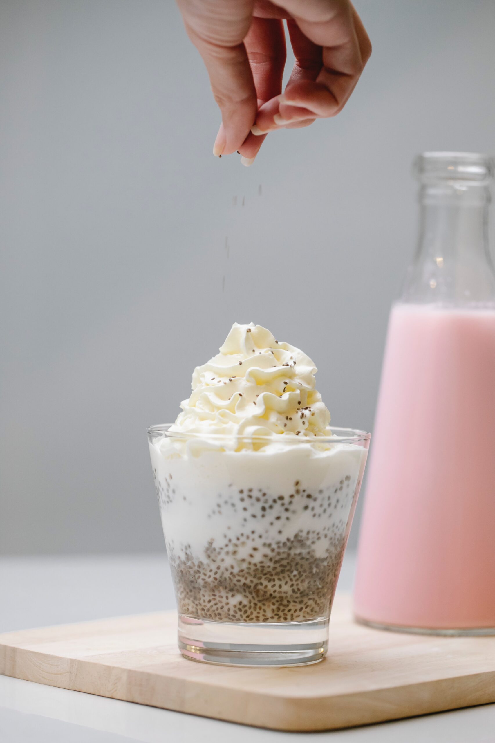 Chia-Pudding im Glas mit Schlagobers, darüber werden Chiasamen gestreut, daneben eine Flasche rosa Milch